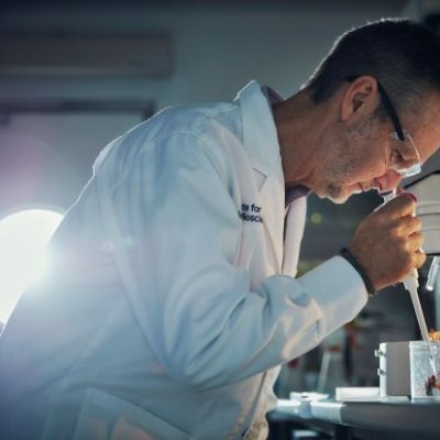 A male scientist in a lab coat and goggles holds a pipette above a large black spider in a container.